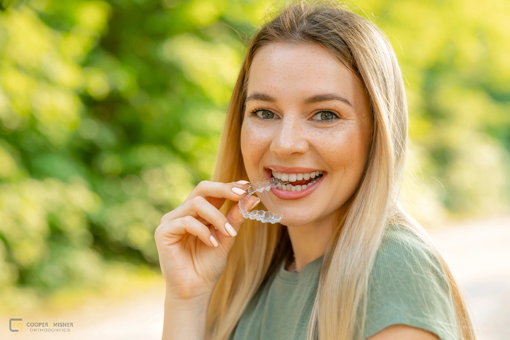 Smiling woman holding a clear aligner outdoors—showing one option for how to fix overbite in Tulsa, OK with Invisalign.
