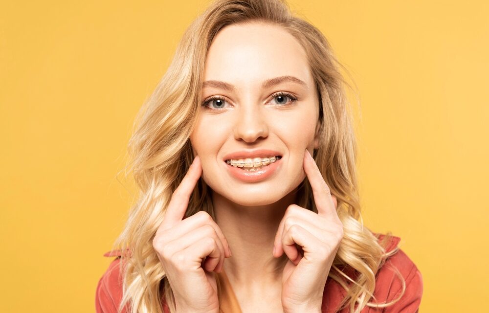 Smiling blonde woman pointing at her teeth against a yellow background - overbite vs underbite in Tulsa, OK.